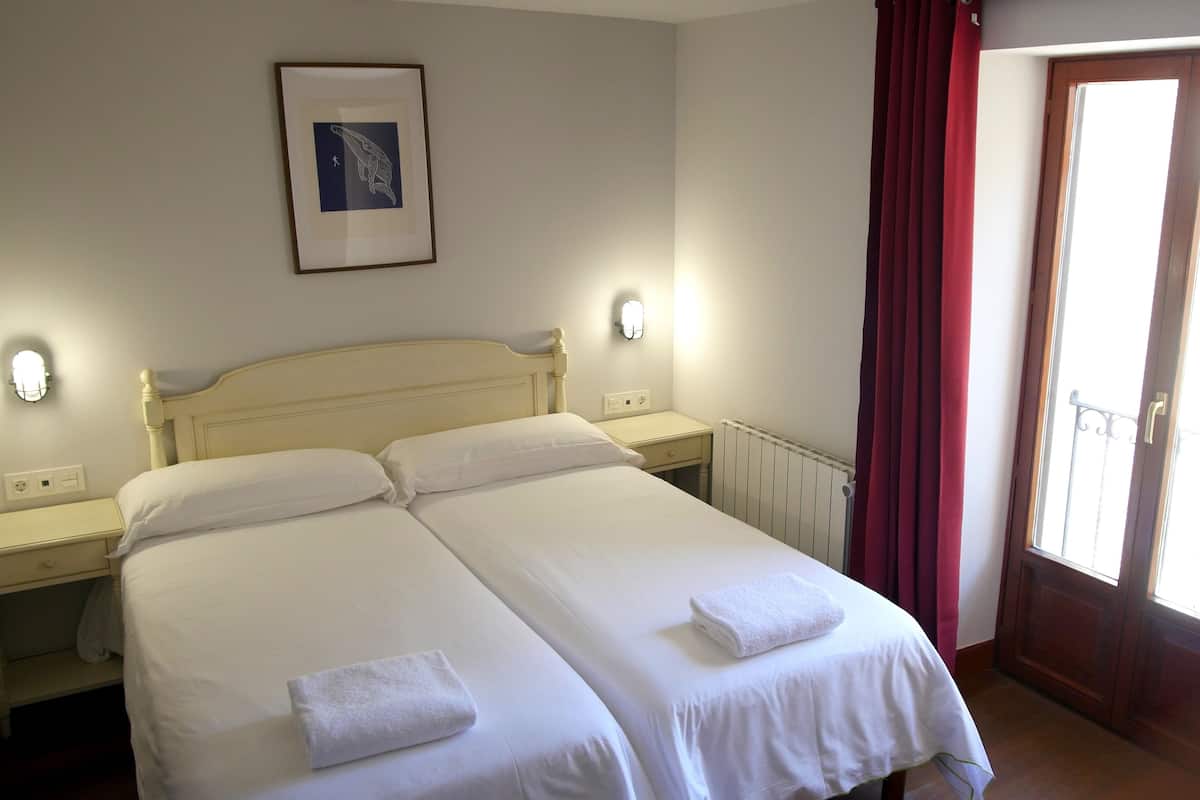 A double bed with white linens and two towels, a bedside table with a lamp, a framed picture on the wall, and a window with red curtains.