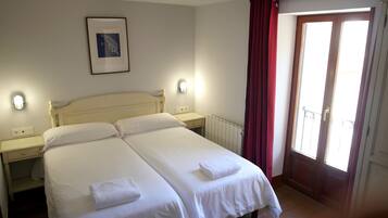 Una cama doble con sábanas blancas y dos toallas, una mesita de noche con lámpara, un cuadro en la pared y una ventana con cortinas rojas.