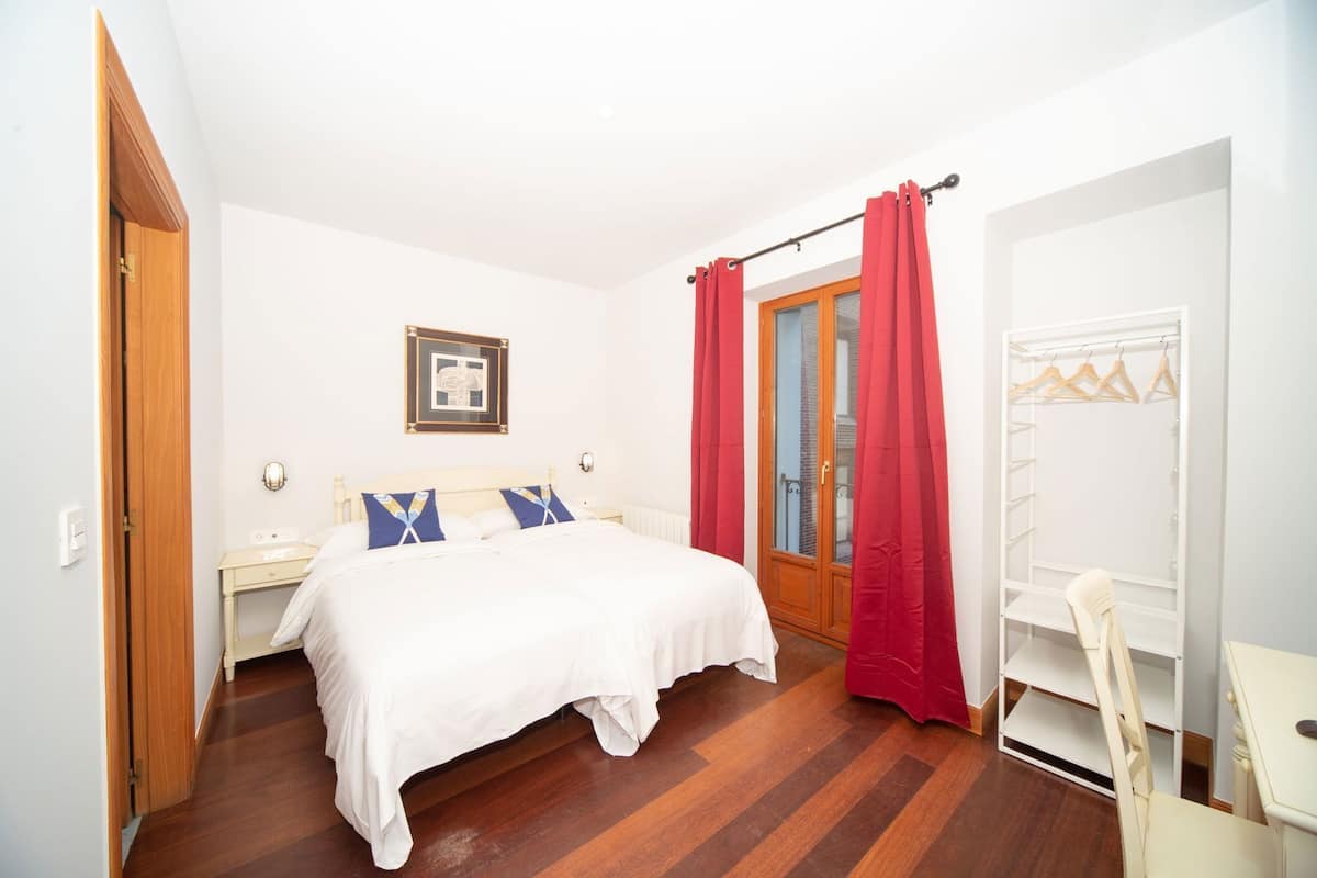 A bedroom with a wooden floor, a bed with white bedding, a nightstand, a wall-mounted light, a door with a red curtain, and a white wardrobe with a chair.