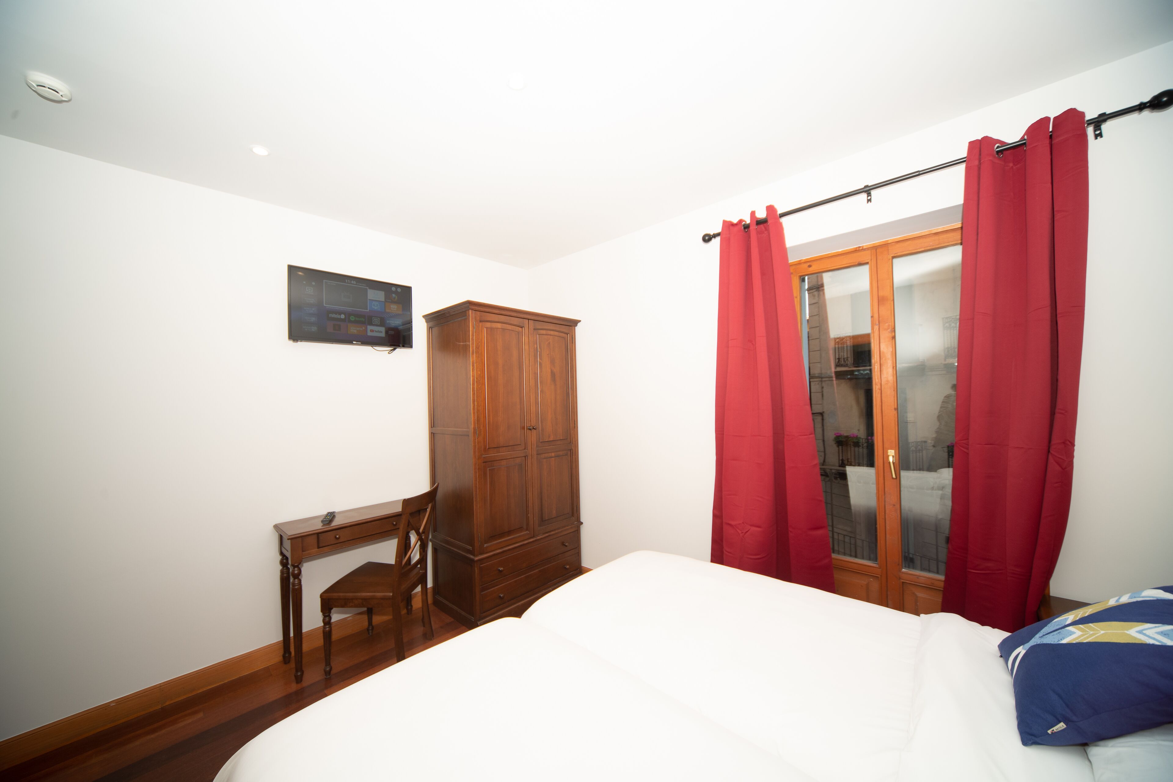 Un dormitorio con una cama, un armario de madera, un escritorio y una ventana con cortinas rojas.