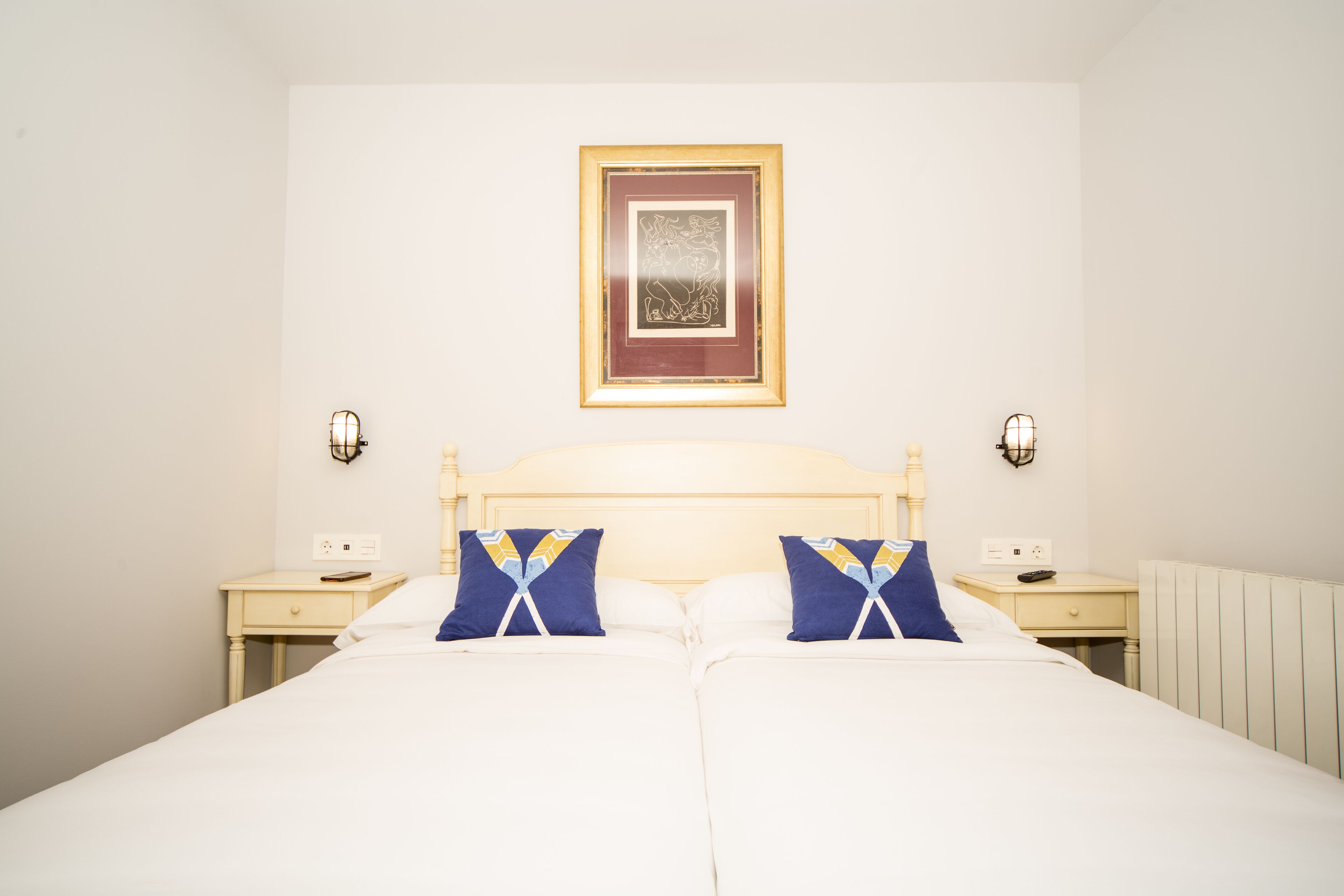 Cama de casal com roupa de cama branca, duas mesas de cabeceira e um quadro na parede.