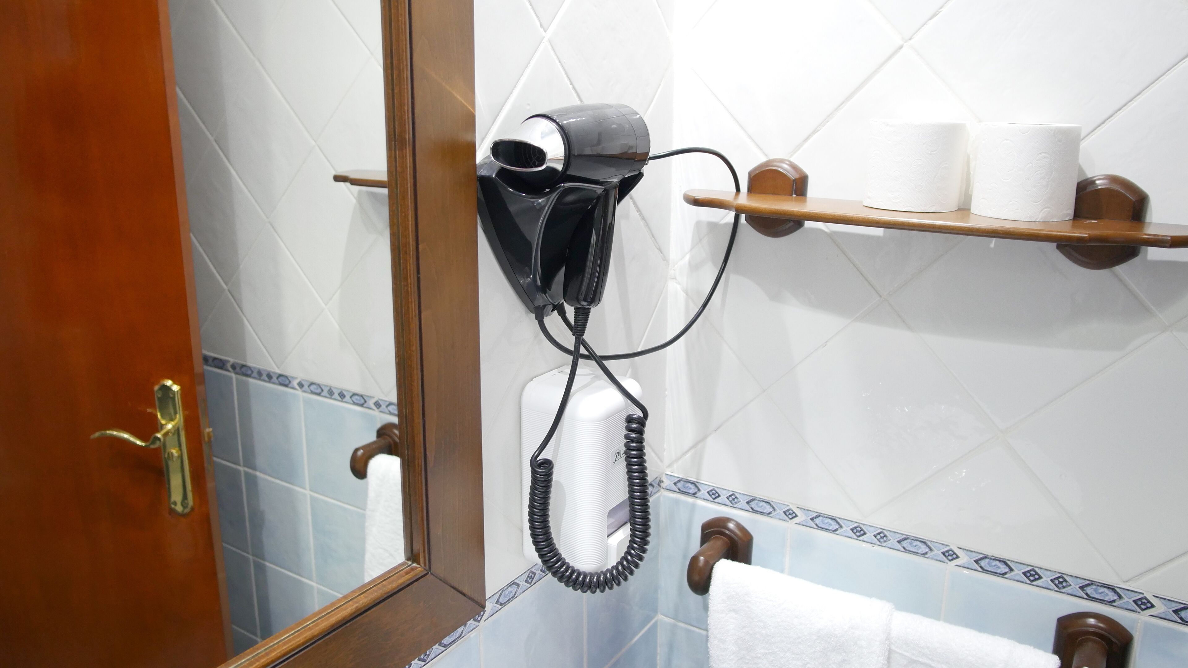 A bathroom with a hairdryer mounted on the wall, a towel rack, and a shelf with toiletries.