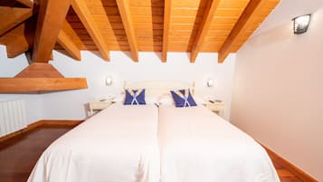 A bedroom with a large bed, wooden ceiling beams, white walls, and a nightstand with a lamp.