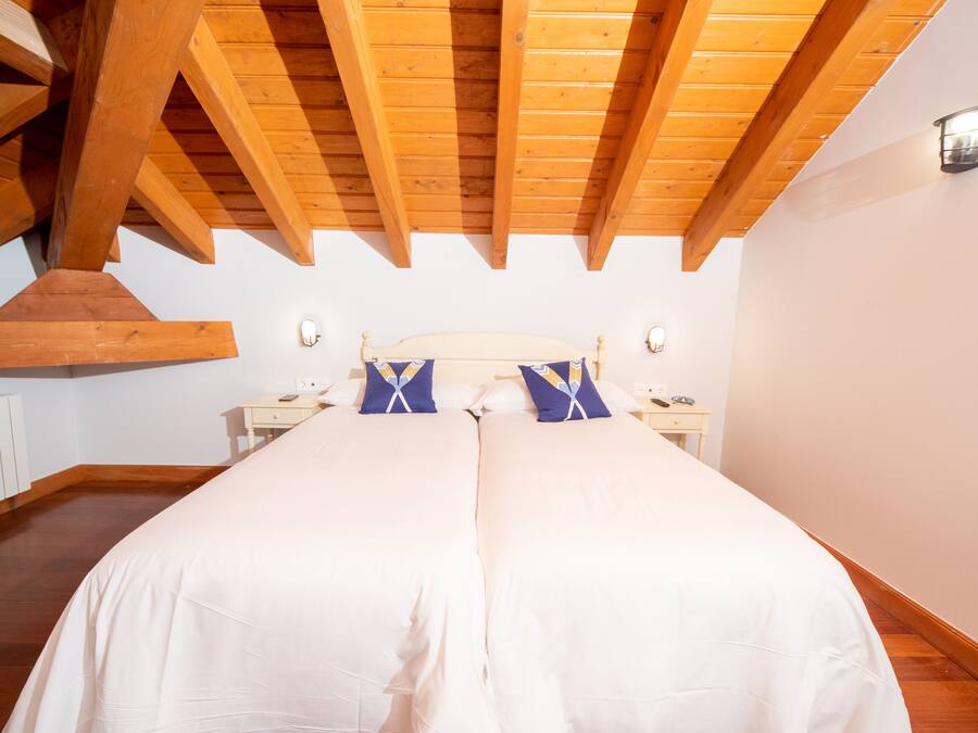 A bedroom with a large bed, wooden ceiling beams, white walls, and a nightstand with a lamp.