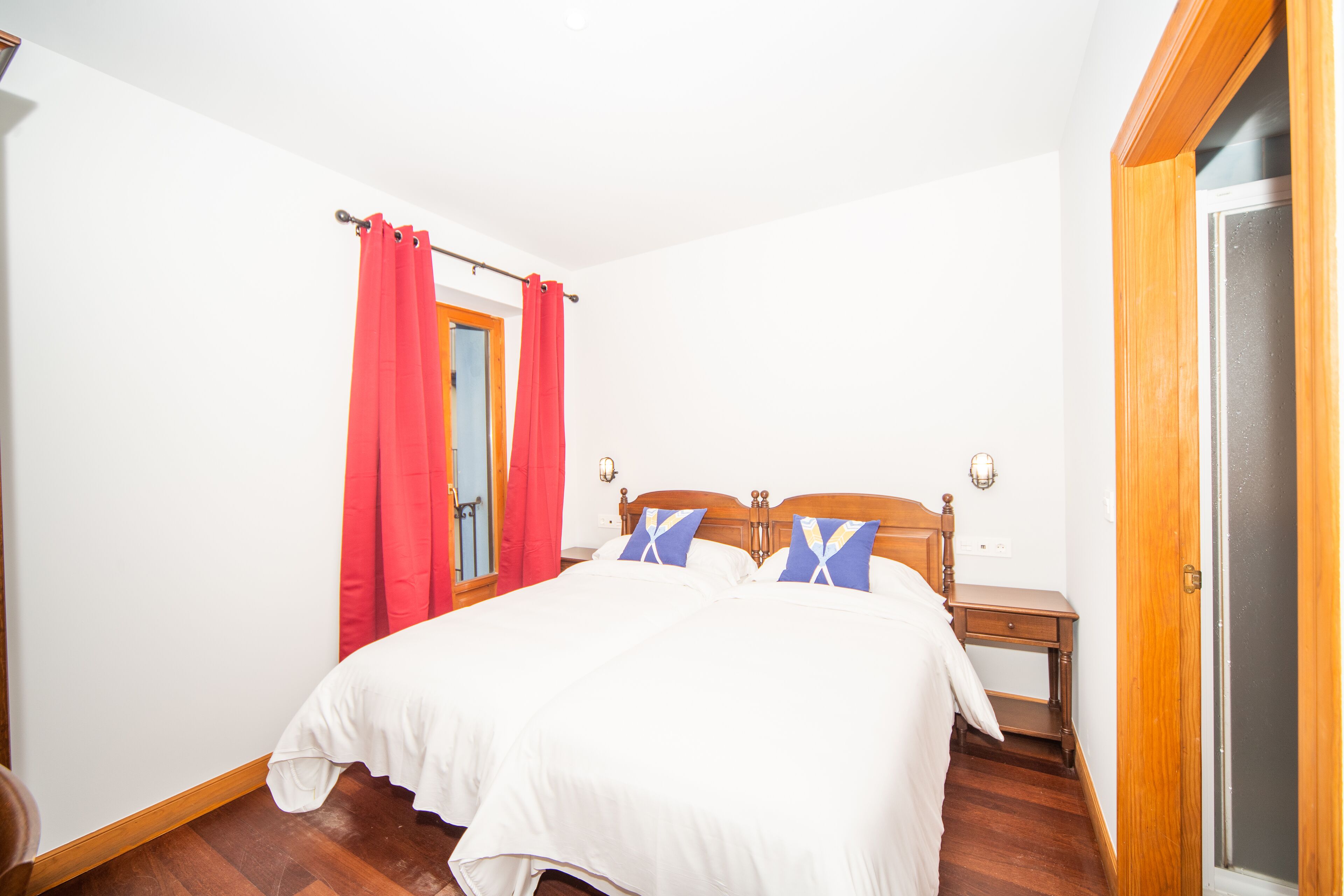 Un dormitorio con cama de madera, dos almohadas, una mesita de noche y una cortina roja.