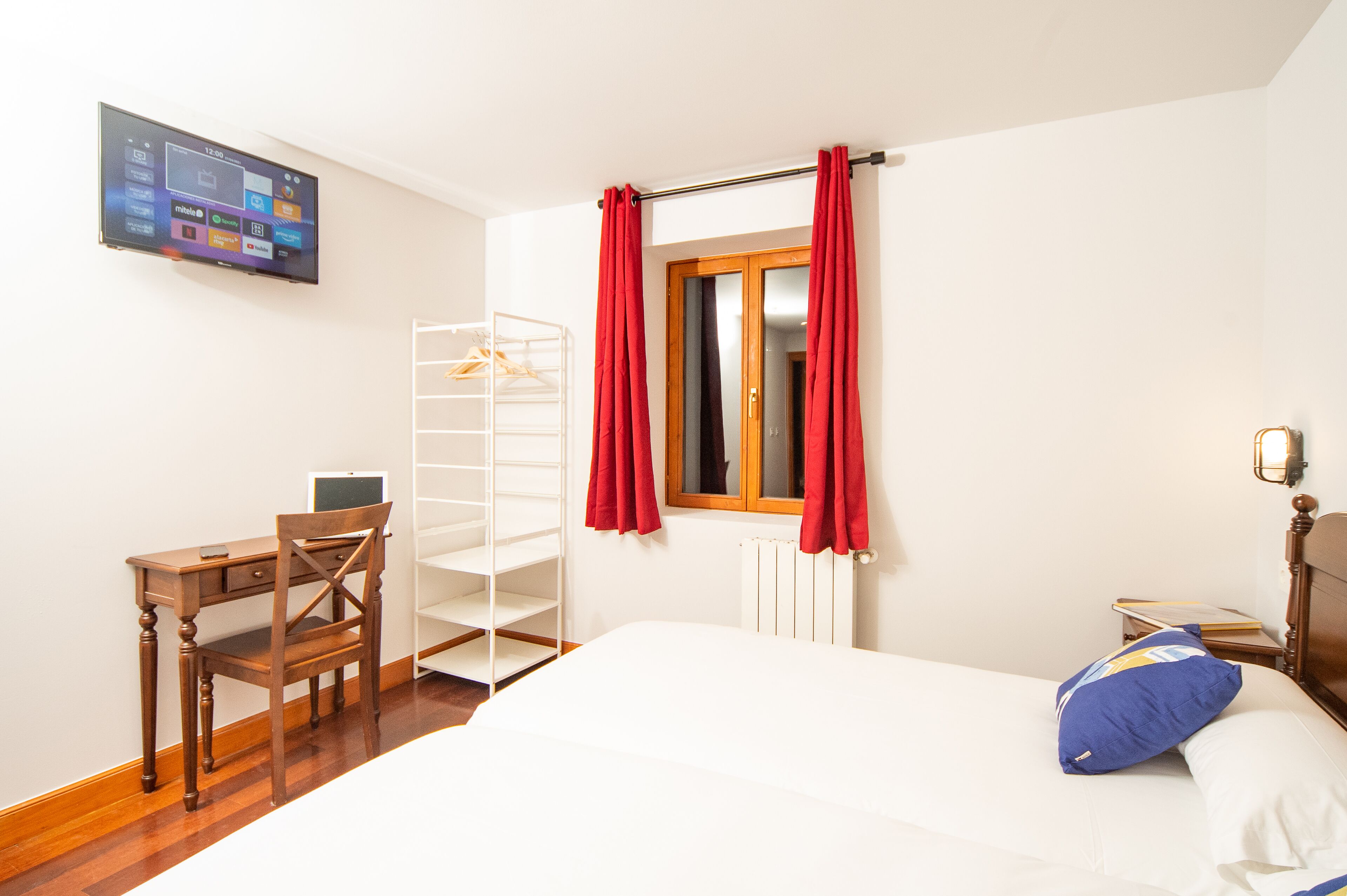 A bedroom with a bed, a desk with a chair, a shelf unit, and a TV mounted on the wall.