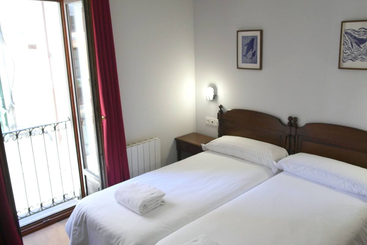 A hotel room with two single beds, a window with red curtains, and two wall-mounted light fixtures.