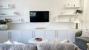 Ein modernes Wohnzimmer mit Flachbildfernseher, weißen Regalen mit Dekoration, einem runden Couchtisch und einem grauen Sofa.