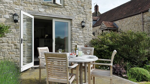 An outdoor dining area with a stone building, a table with chairs, and a glass door leading to an interior space. - The Barn, Park Farm near Chew Magna, Bristol (Bristol)
