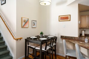 Un coin repas avec une table en bois foncé, dressée pour quatre personnes, entourée de chaises dotées de coussins de couleur claire. Des œuvres d’art encadrées ornent les murs, et un escalier avec une rampe en bois est visible sur la gauche.