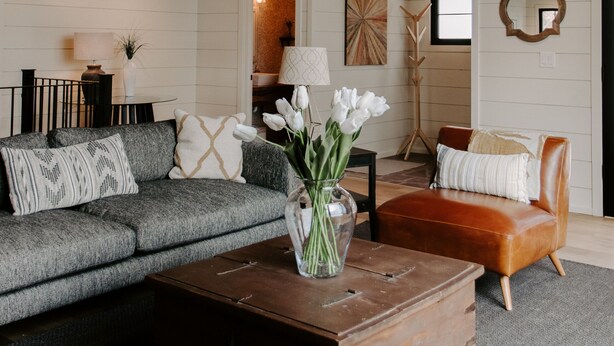 Ein gemütliches Wohnzimmer mit Holzbalkendecke, einem grauen Moduls Sofa, einem braunen Ledersessel und einem Holztisch mit Blumenvase.