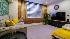 Un salon moderne avec un canapé gris, des coussins jaunes, une table basse en verre, un téléviseur à écran plat et un mur en briques.