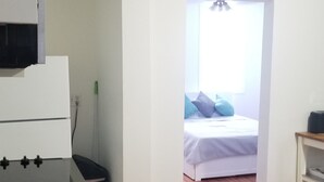 A kitchen with a black countertop and a white stove. There is a wooden floor and a doorway leading to a bedroom with a bed and a window. - Spacious Studio near downtown (Newport)