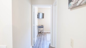 Ein Flur mit Holzboden, einer weißen Tür, einem kleinen Tisch mit Mülleimer und einem Wand-Fernseher.