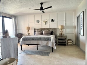 Une chambre à coucher comprenant un lit, un ventilateur de plafond, des lampes de chevet, des éléments décoratifs sur les murs et une vue sur l’extérieur.