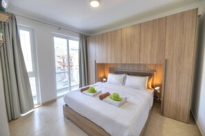 A hotel room with a large bed, wooden headboard, and a view of buildings through the window. - Beautifully furnished 2 bedroom apartment DDIF1-3 (Gzira)