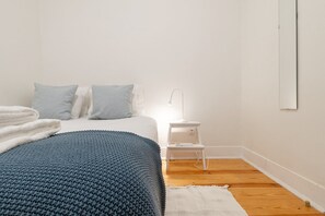 A bedroom with a bed, pillows, a bedside table with a lamp, a white rug, and a wooden floor.