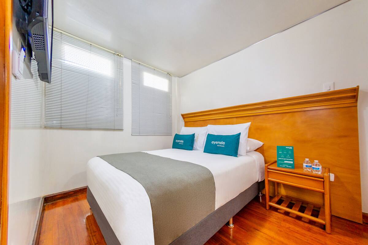 Une chambre d’hôtel avec un lit, une table de chevet en bois sur laquelle se trouve une bouteille d’eau, et une enseigne indiquant le nom de l’hôtel.