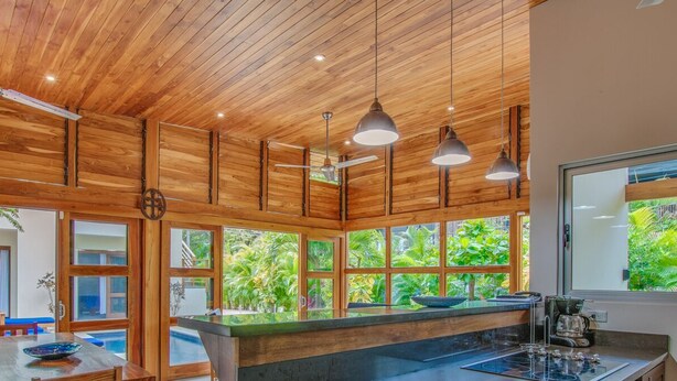 Une cuisine moderne dotée d’un grand îlot central, d’un plafond en bois et de grandes fenêtres donnant sur un jardin luxuriant.