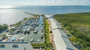 Eine Luftaufnahme einer Marina mit vertäuten Booten, ein längeres Gebäude mit flachem Dach und freier Sicht auf den Ozean und eine entfernte Insel.