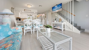 Ein modernes Wohnzimmer mit einem Glastisch, weißen Korbstühlen, einer Couch mit blau-korallfarbenem Muster und einer Treppe mit Wandkunst in Blau.