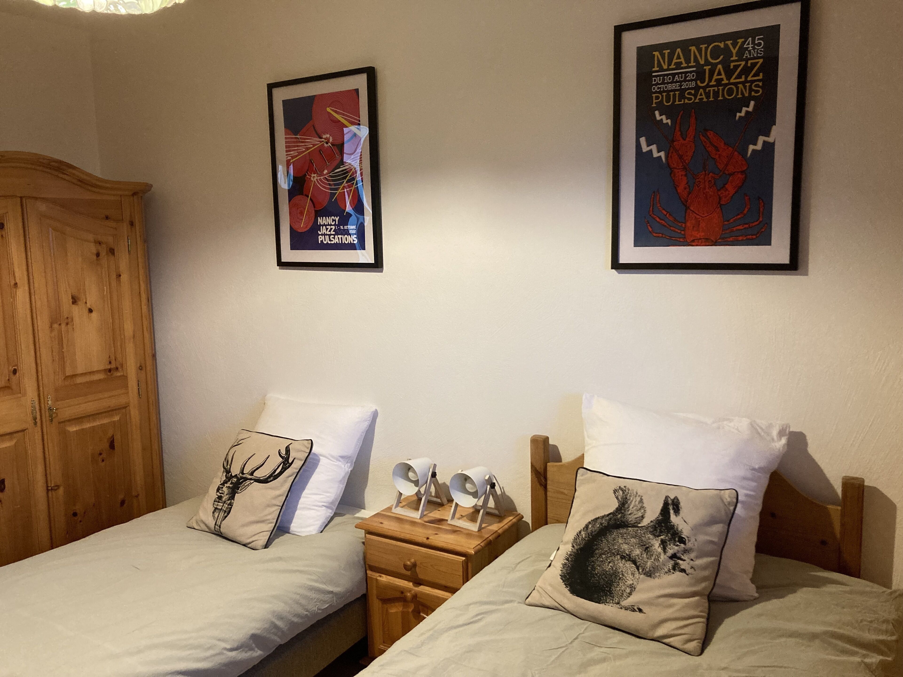 Une chambre avec deux lits, chacun orné d’un coussin à motifs représentant un cerf et un raton laveur. Des affiches encadrées ornent le mur, dont l’une est intitulée « Nancy Jazz Pulsations ». Une armoire en bois est visible sur la gauche.
