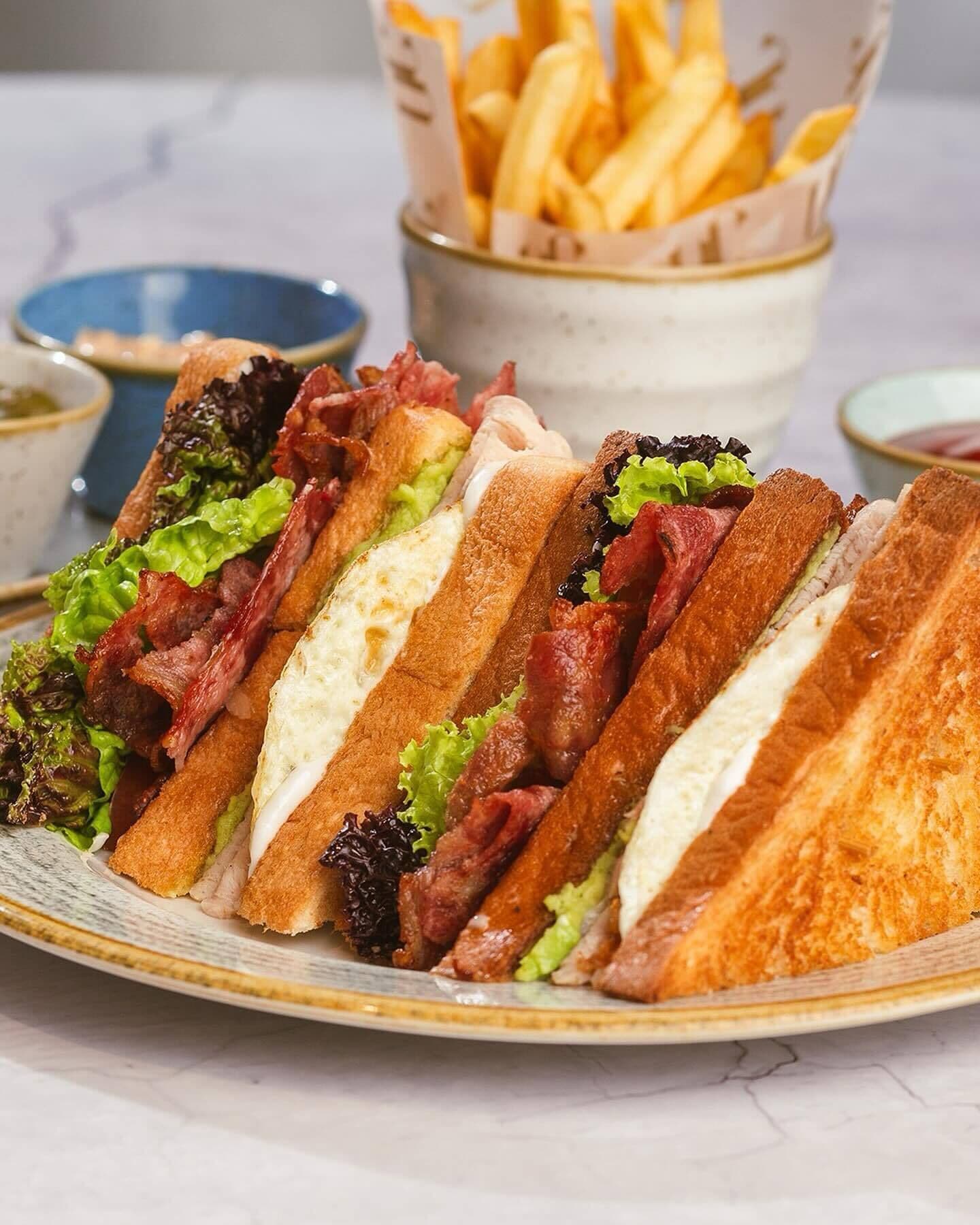 Ein Teller mit Sandwiches, belegt mit Salat, Speck und Käse, dazu ein Korb Pommes und eine kleine Schale Sauce.