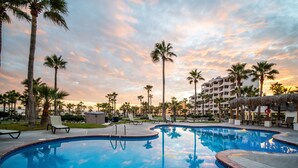 A swimming pool with lounge chairs, palm trees, and a hotel building in the background.