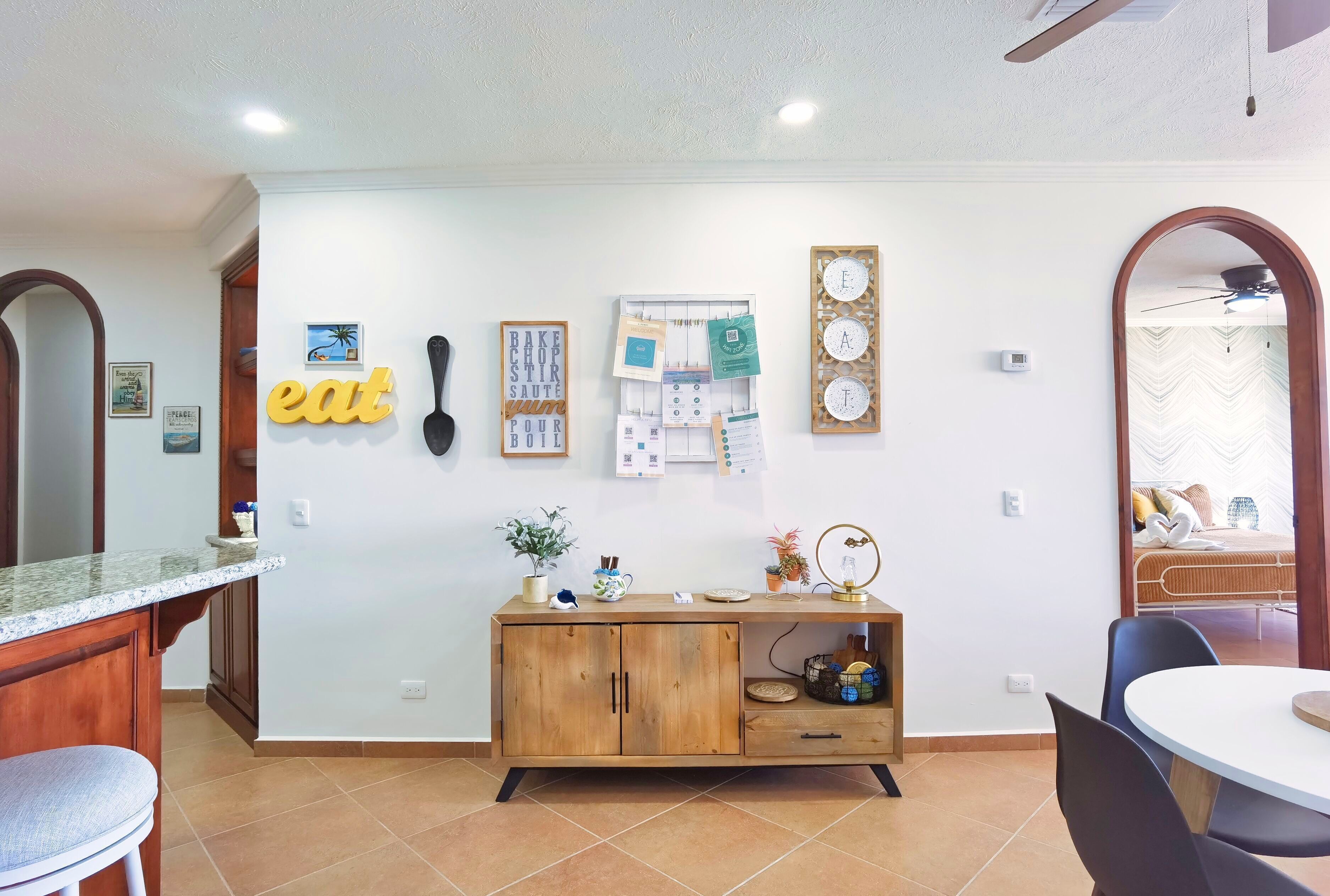 A modern kitchen with a wooden cabinet, a dining area with a round table, and a wall with various decorations including a clock and a bulletin board.