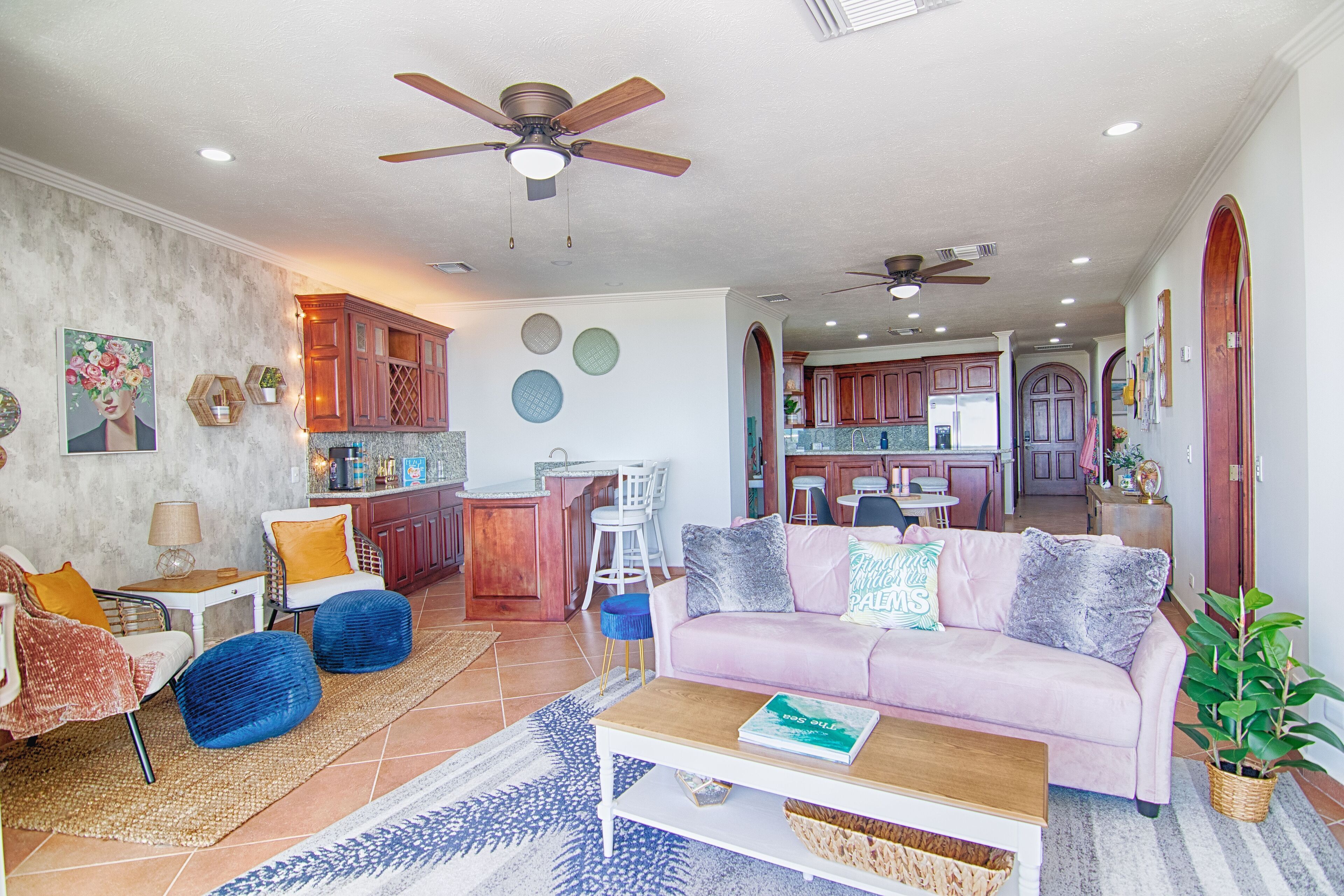 A spacious living area with a pink sofa, blue chairs, and a coffee table. There is a kitchen in the background with wooden cabinets and a dining area.