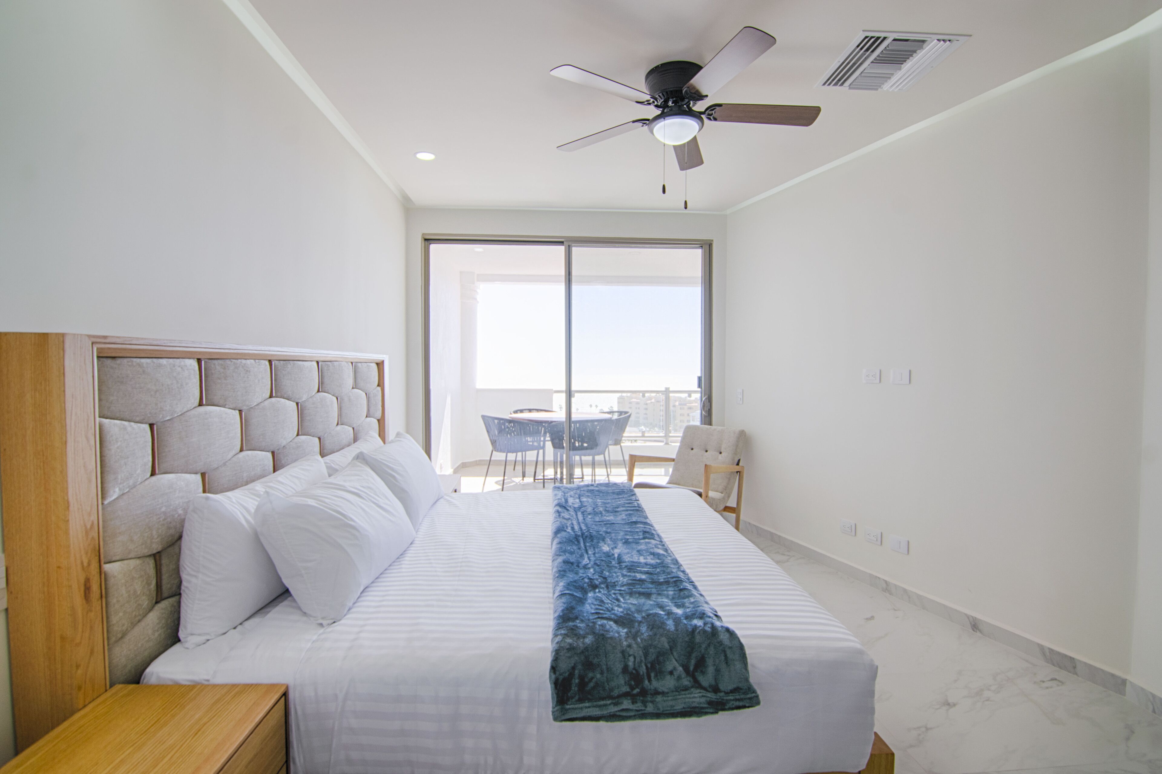 A hotel room with a large bed, a ceiling fan, a view of the outdoors, a small table, and a chair.