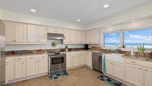 A modern kitchen with white cabinets, granite countertops, a stainless steel oven, and a view of the ocean through large windows. - Shore Beats Work Home, Deck & Oceanfront Views (Ponte Vedra Beach)