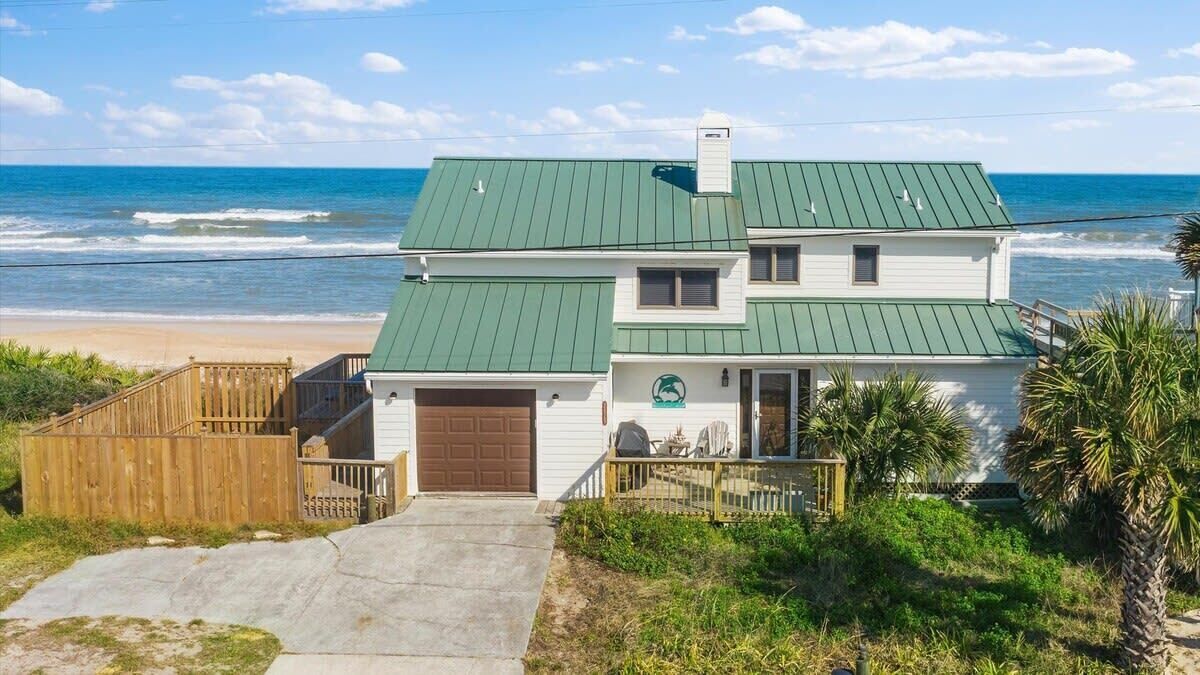 Uma casa de dois andares com telhado verde, varanda frontal e vista para a praia.
