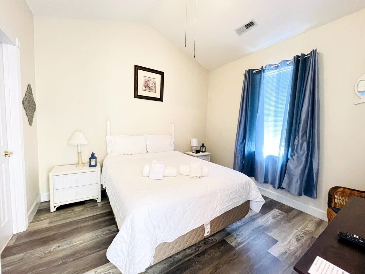 Una camera d'albergo con un letto grande, un comodino, una lampada, un quadro appeso al muro e una finestra con tende.