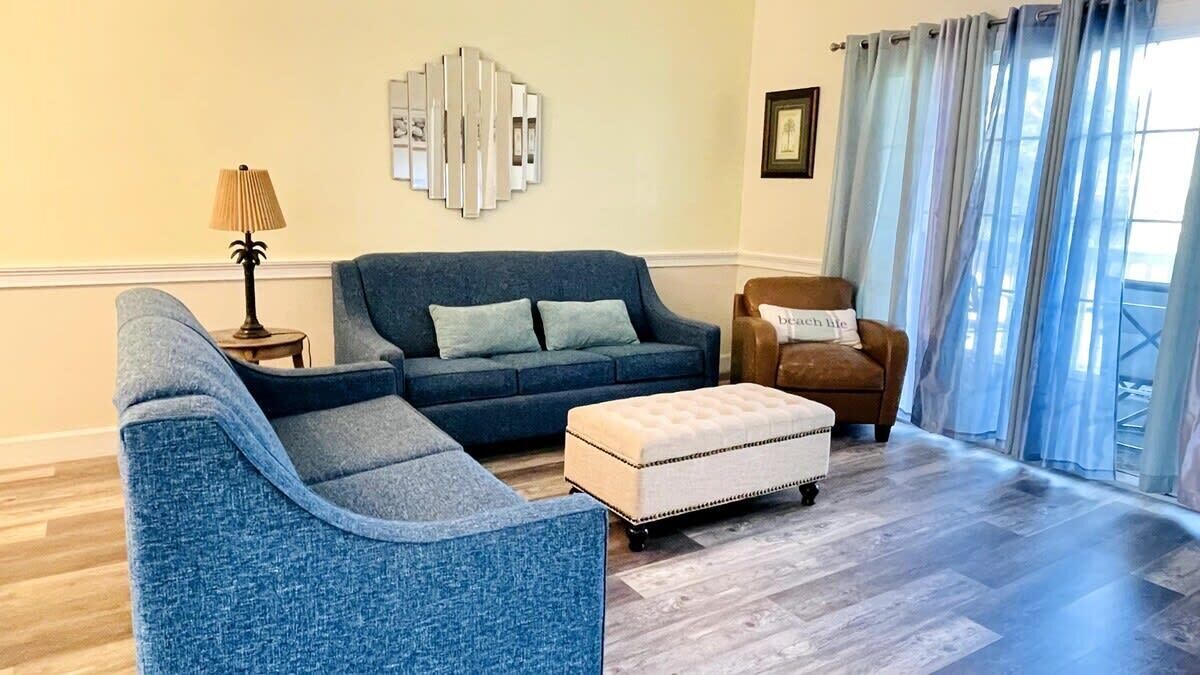A living room with a blue sofa, a matching armchair, a small ottoman, a lamp, and a wall art piece.