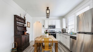 Une cuisine avec une table à manger en bois, une chaise jaune, des placards blancs, un réfrigérateur en acier inoxydable et un buffet en bois foncé.