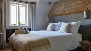 Ein ordentlich bezogenes Bett mit weißer Bettdecke und Kissen, eine Nachttischlampe, ein Fenster mit Vorhängen und eine Heizung.
