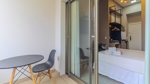 A hotel room with a sliding glass door leading to a balcony, a bed with neatly folded towels, a small table with two chairs, and a wardrobe with hanging clothes. - Studios E-side Vila Madalena (São Paulo)
