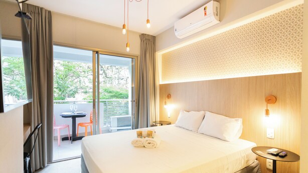A hotel room with a large bed, a small table, a chair, and a balcony with a table and chairs. - Studios E-side Vila Madalena (São Paulo)