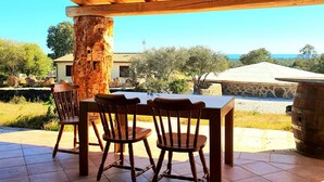 Eine Terrasse im Freien mit einem Holztisch und -stühlen, einer Steinsäule und Blick auf das Meer sowie Gebäude im Hintergrund.