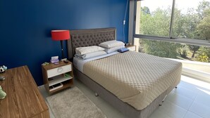A bedroom with a bed, a nightstand, a desk, and a window with a view of greenery. - Donna Alda Tower incredible views (Salta)