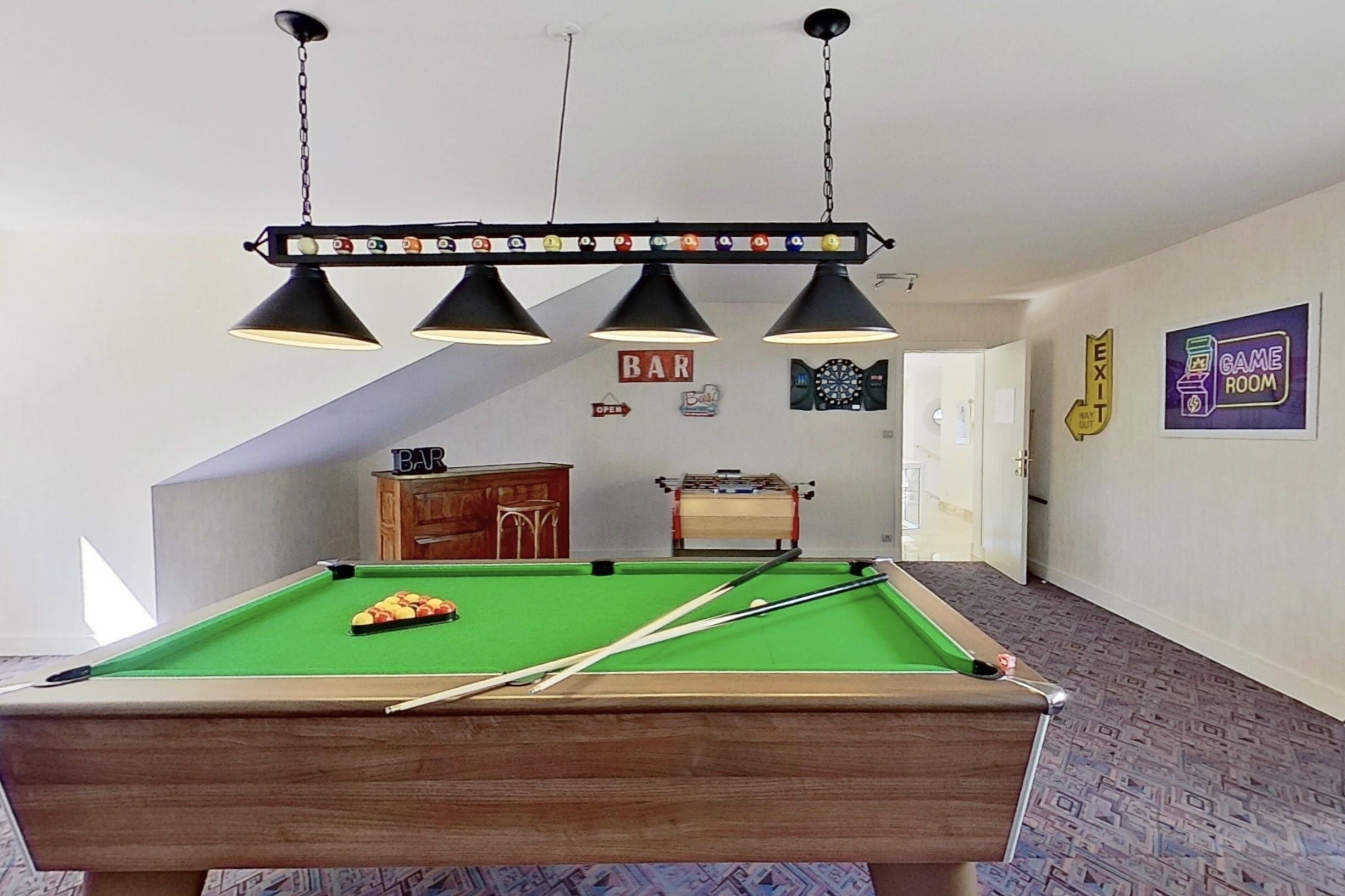 A pool table with a green felt surface, three black pendant lights, and a 'BAR' sign on the wall.