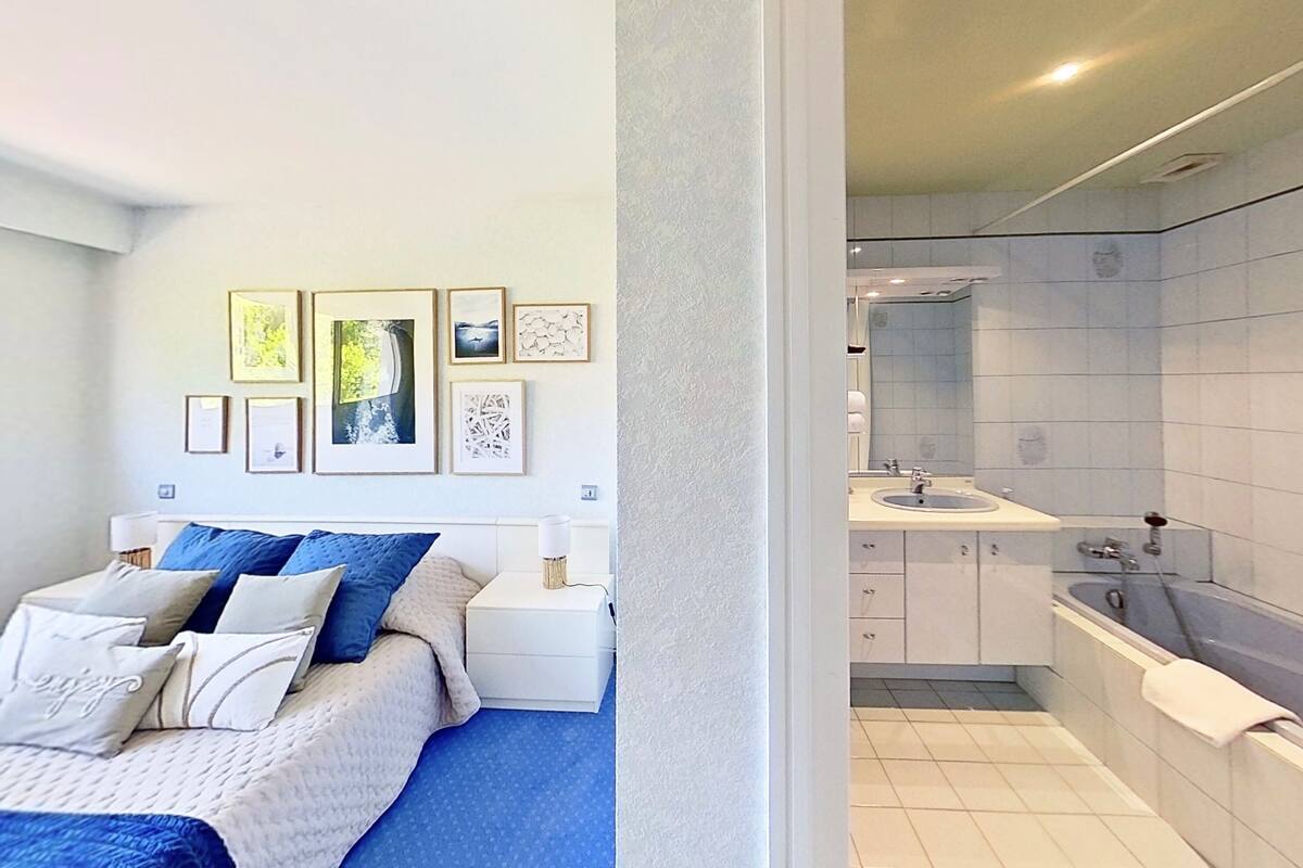 A bedroom with a bed, bedside tables, and wall art. Adjacent is a bathroom with a sink, mirror, and bathtub.