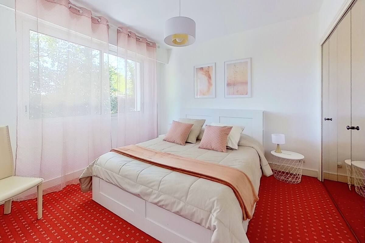 A bedroom with a bed, a chair, a bedside table with a lamp, and a window with curtains.