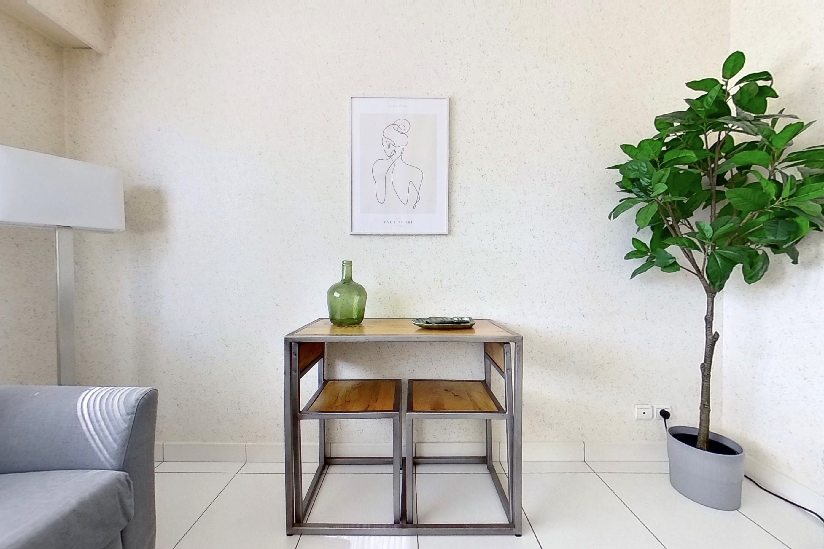 Ein Zimmer mit einem Tisch aus Metallgestell, einer Topfpflanze, einem gerahmten Kunstwerk und einem grauen Sofa.