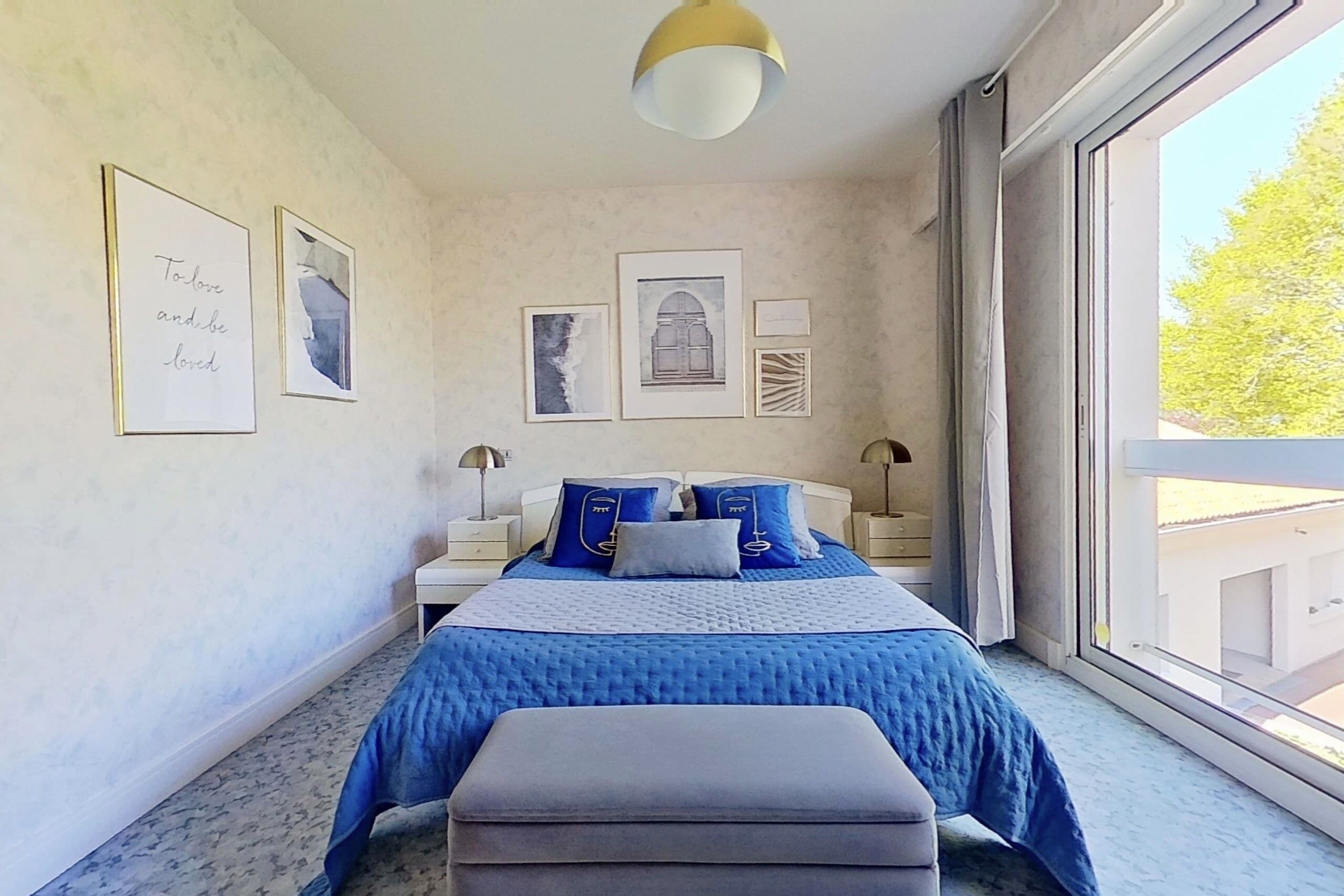Un dormitorio con un ventanal grande, una cama con ropa de cama azul y obras de arte enmarcadas en la pared.