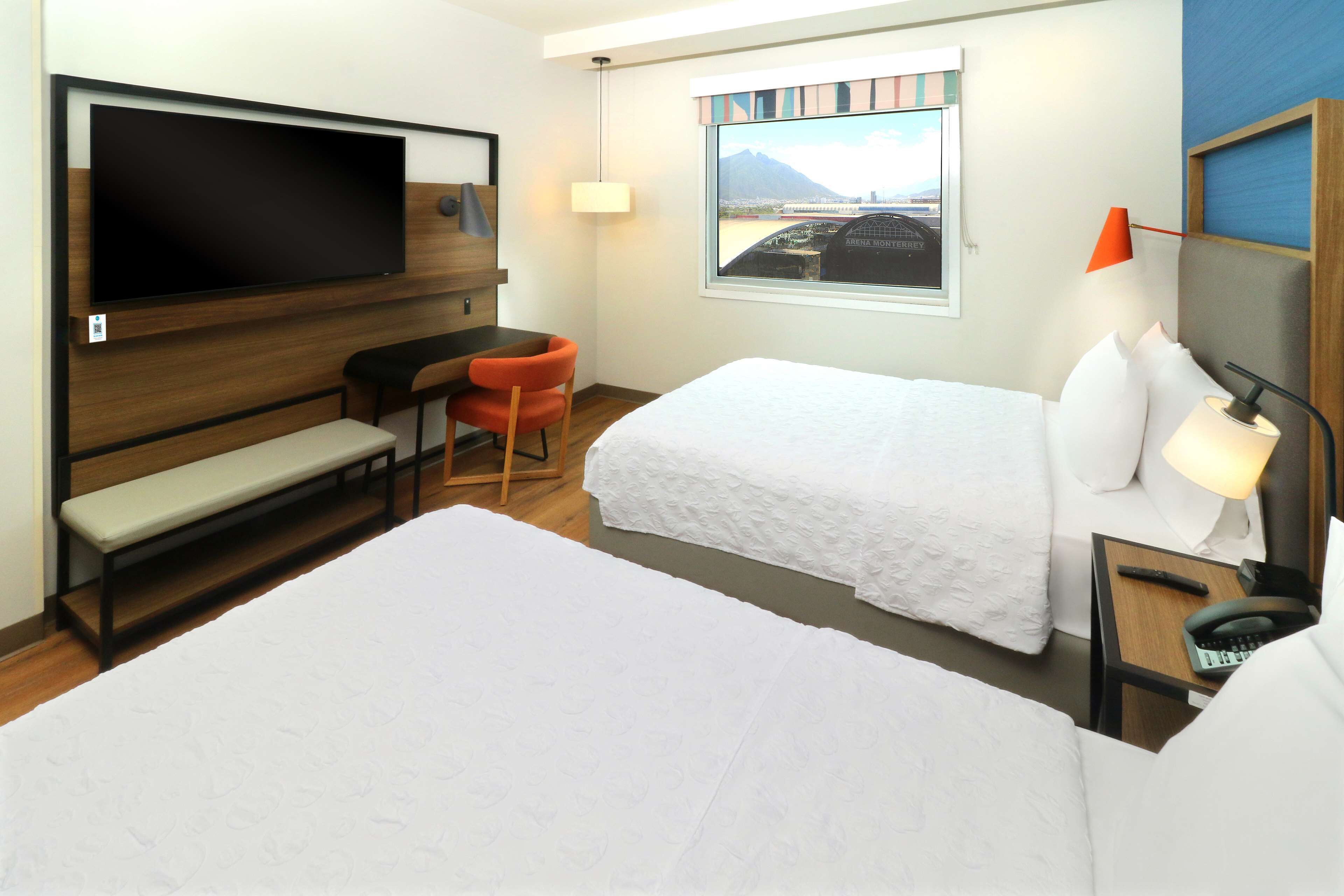 Habitación de hotel con una cama grande, una cama más pequeña, un escritorio con una silla, un televisor montado en la pared, una ventana con vistas a una montaña y una lámpara en la mesita de noche.