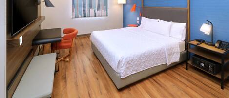 A hotel room with a bed, a desk, a chair, a TV, and a painting on the wall.