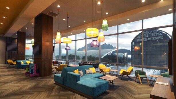 A modern lounge area with a large sofa, colorful hanging lamps, and a view of a bridge and mountains through large windows. - Tru By Hilton Monterrey Fundidora (Monterrey)