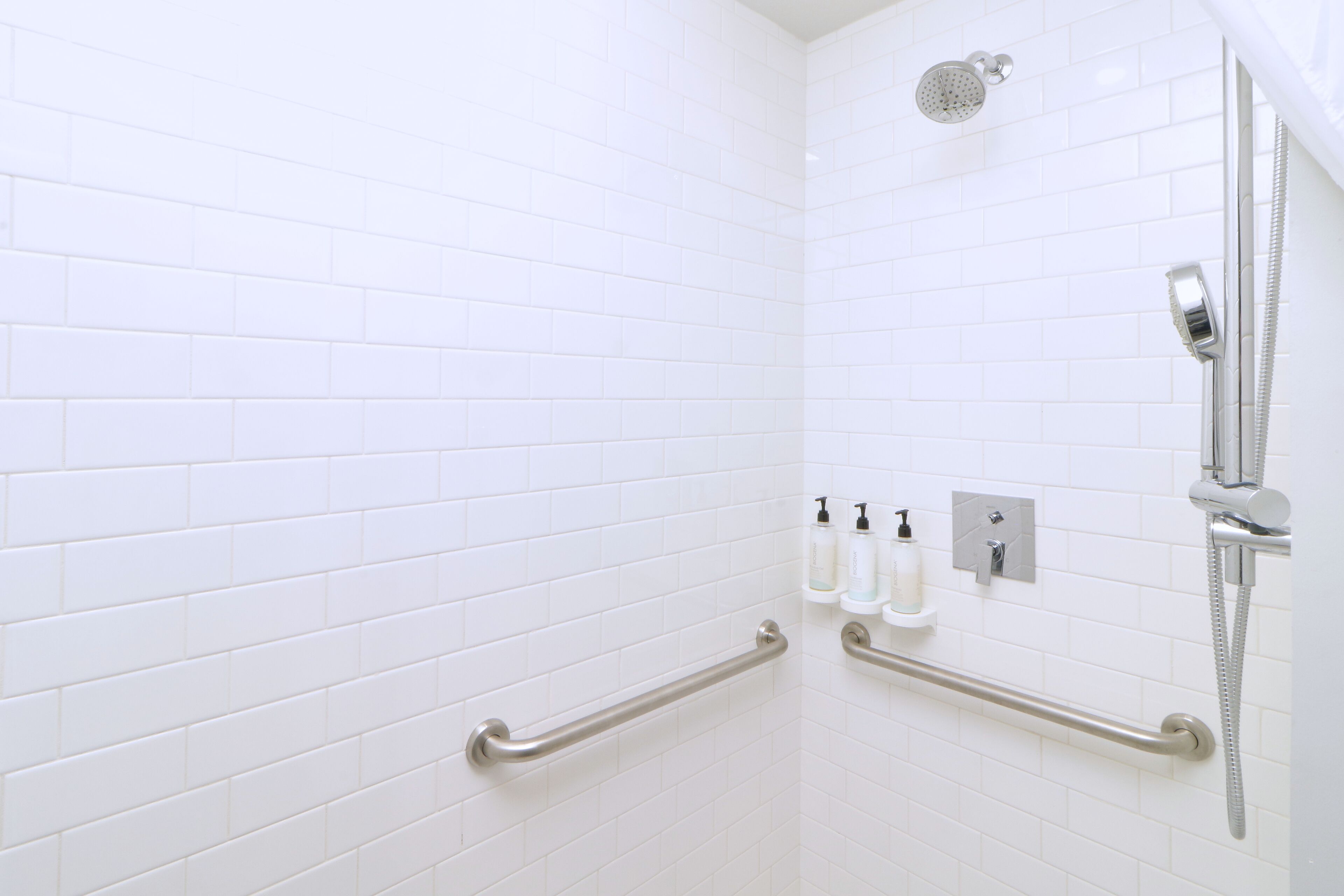 A white-tiled shower area with a handheld showerhead, a handheld showerhead, and a wall-mounted showerhead. There are two grab bars installed for support.